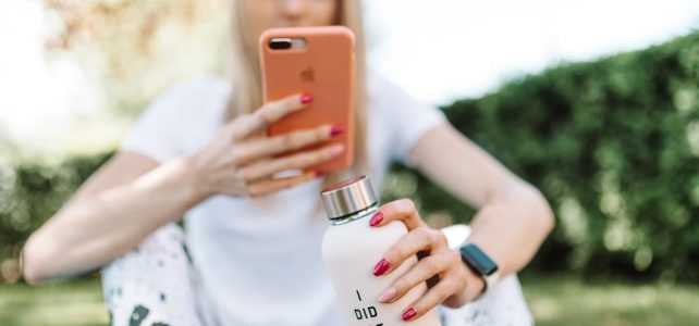 Jak znaleźć motywację do zmiany nawyków Woman outdoors using smartphone while holding a water bottle with text. Casual and relaxed setting.